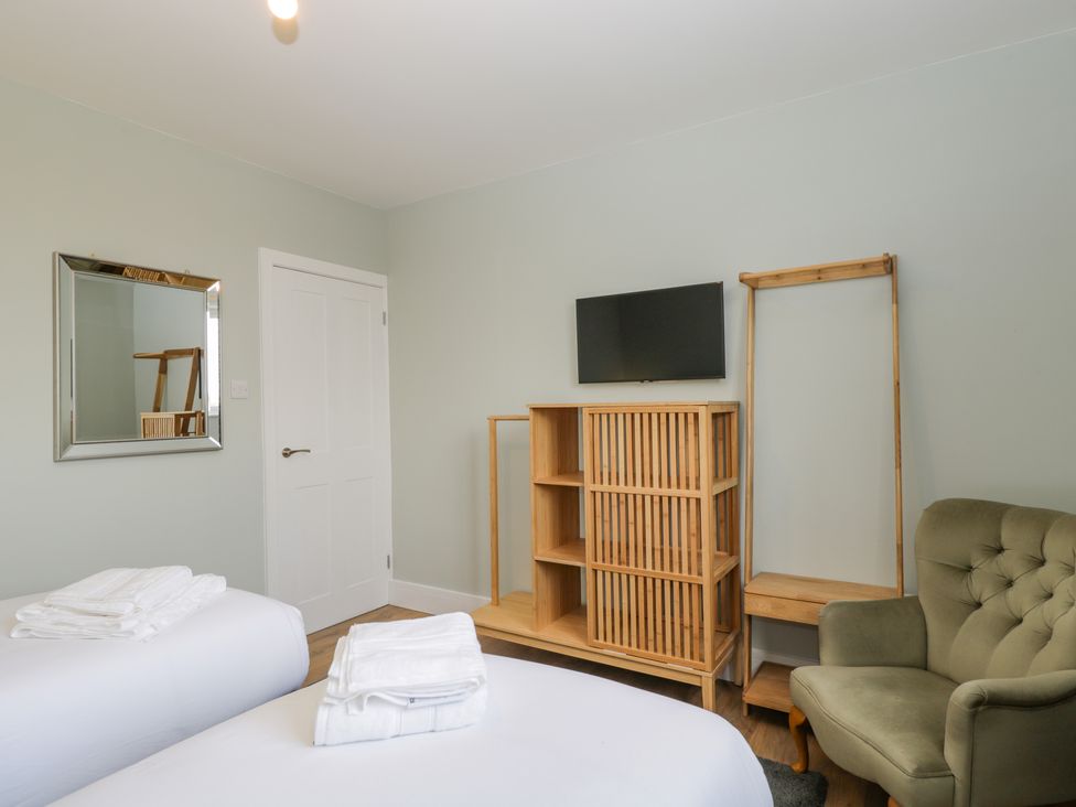 A bedroom with two beds, folded white towels on each, a wooden cabinet, a mounted television, a green armchair, a full-length mirror, and a white door at Birch House in Corsham