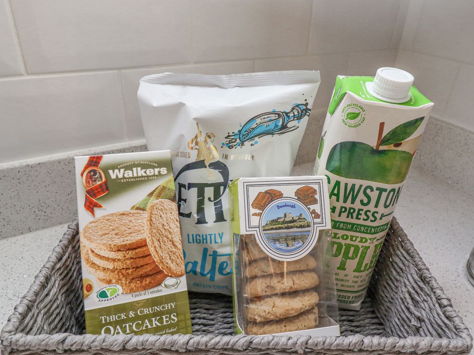 A wicker basket with oatcakes crackers potato chips biscuits and a carton of cloudy apple juice on a kitchen countertop at Coral Cottage in Bamburgh
