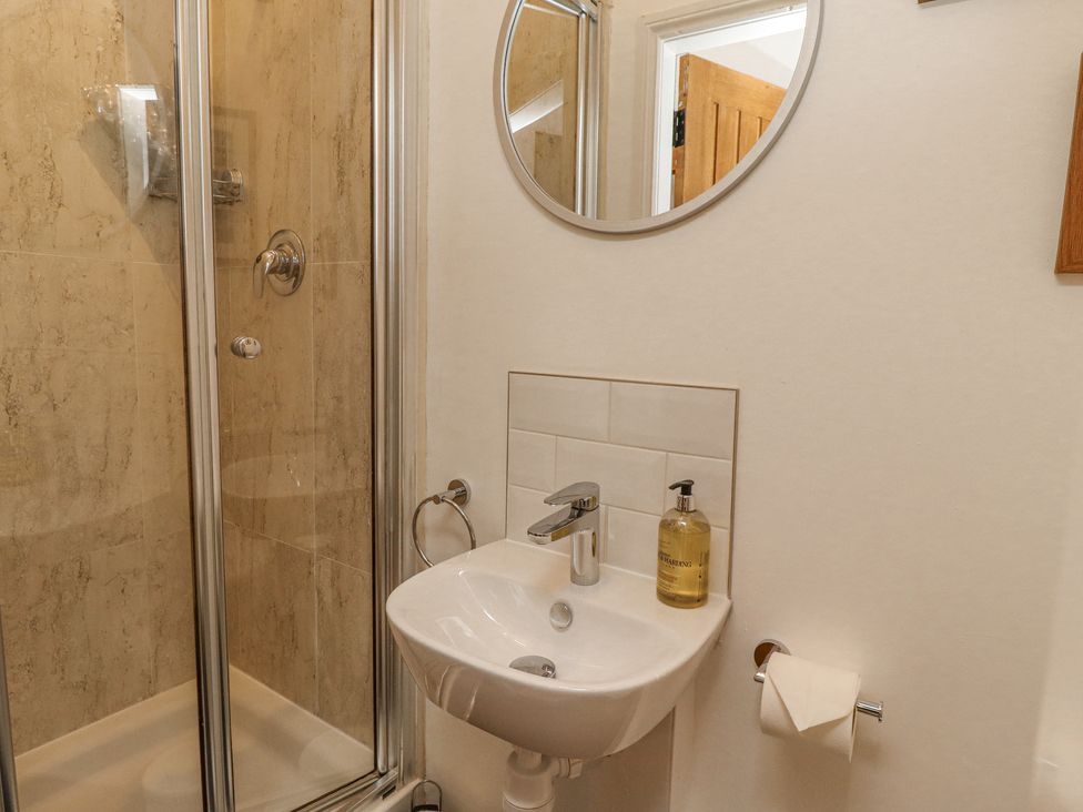 A bathroom with a corner shower, a white sink, a round mirror, and a soap dispenser at Coral Cottage in Bamburgh
