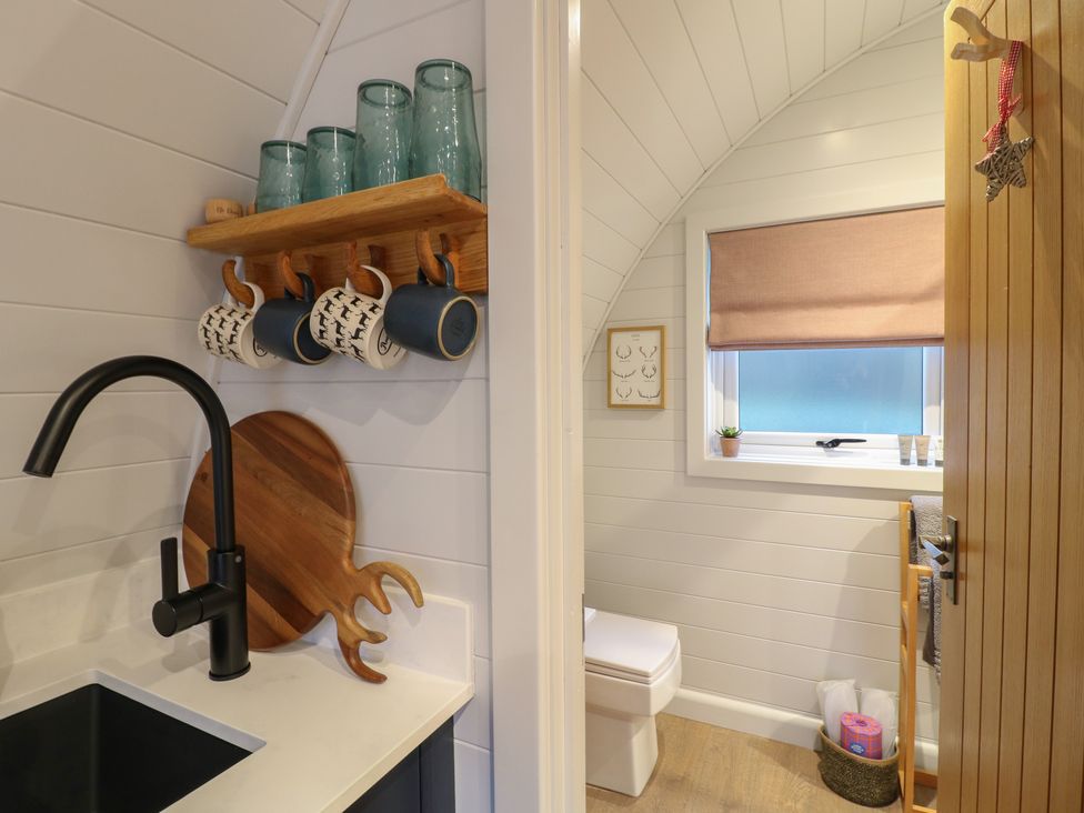 A kitchen sink with black faucet mugs on hooks above and a bathroom with toilet wooden door and window at No Deer in Ashbourne