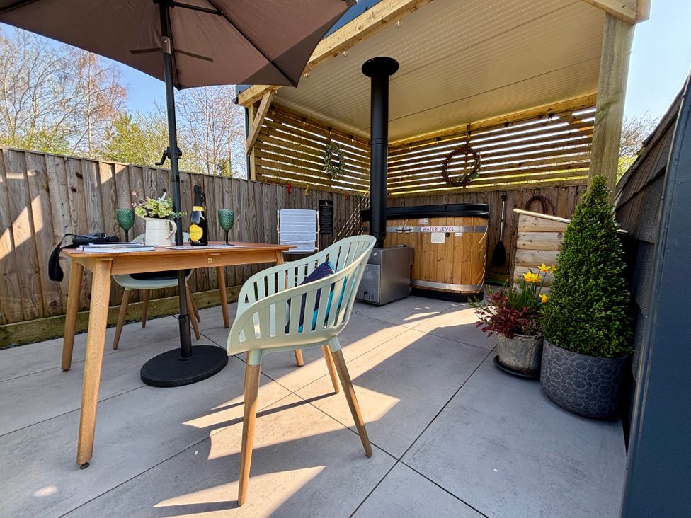 An outdoor patio with a table under an umbrella two chairs a wooden hot tub plants and a wood stove at No Deer in Ashbourne
