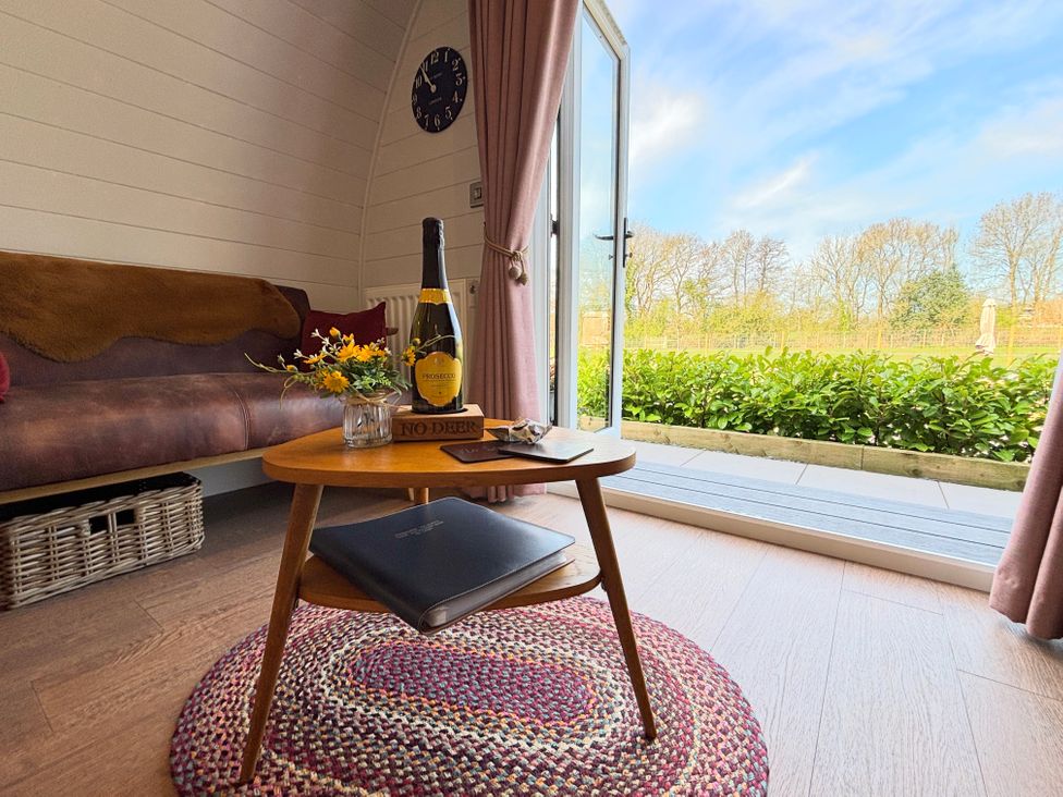 A living area with a wooden coffee table holding a bottle and a small vase of flowers next to a brown sofa and an open door showing greenery outside at No Deer in Ashbourne