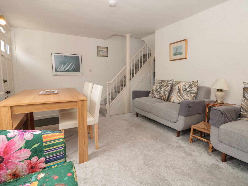 A living room with a wooden dining table and white chairs, gray sofas with patterned cushions, a floral armchair, and a staircase at Dart Cottage in Dittisham