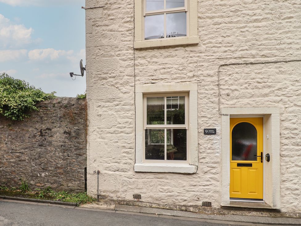 Exterior of a white stone cottage with a yellow door and two windows next to a stone wall at Ribble Cottage in Settle