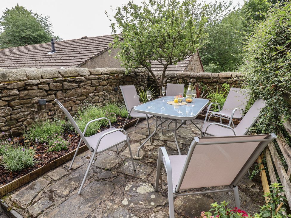 An outdoor patio area with a glass table and six chairs beside a stone wall and plants at Ribble Cottage in Settle