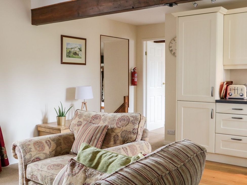 A living area with patterned armchairs a wooden side table with a lamp and plant a kitchen area with cream cabinets and a toaster at The Hayloft at Tennant Barn Malham