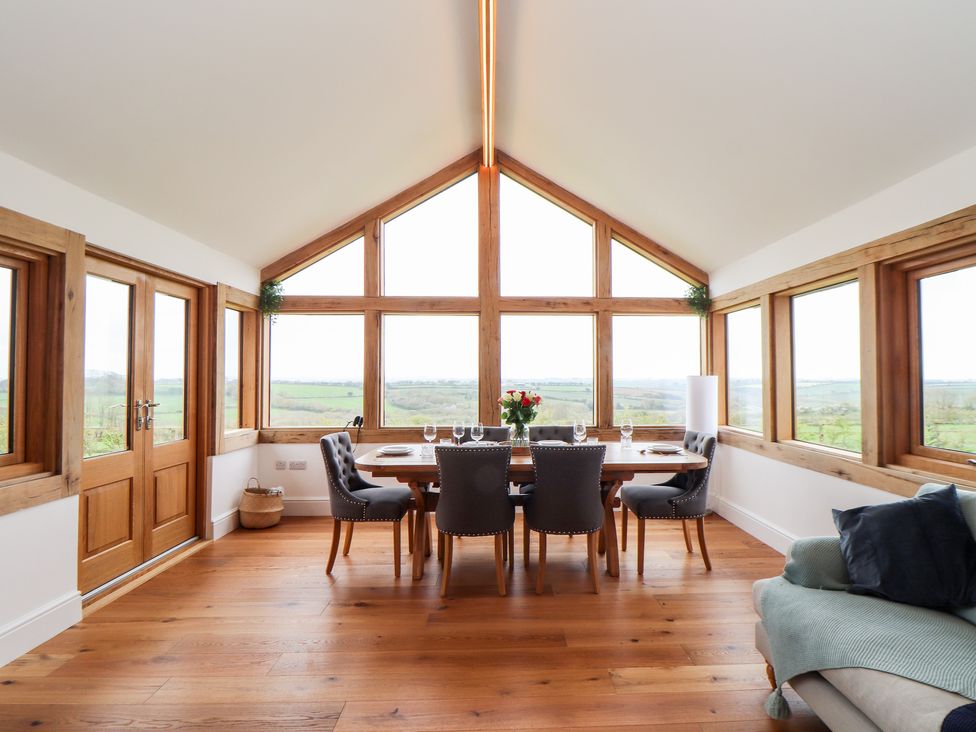 A dining room with wooden table and chairs and large windows showing countryside view at Trevilges Cottage in Trewennack near Helston