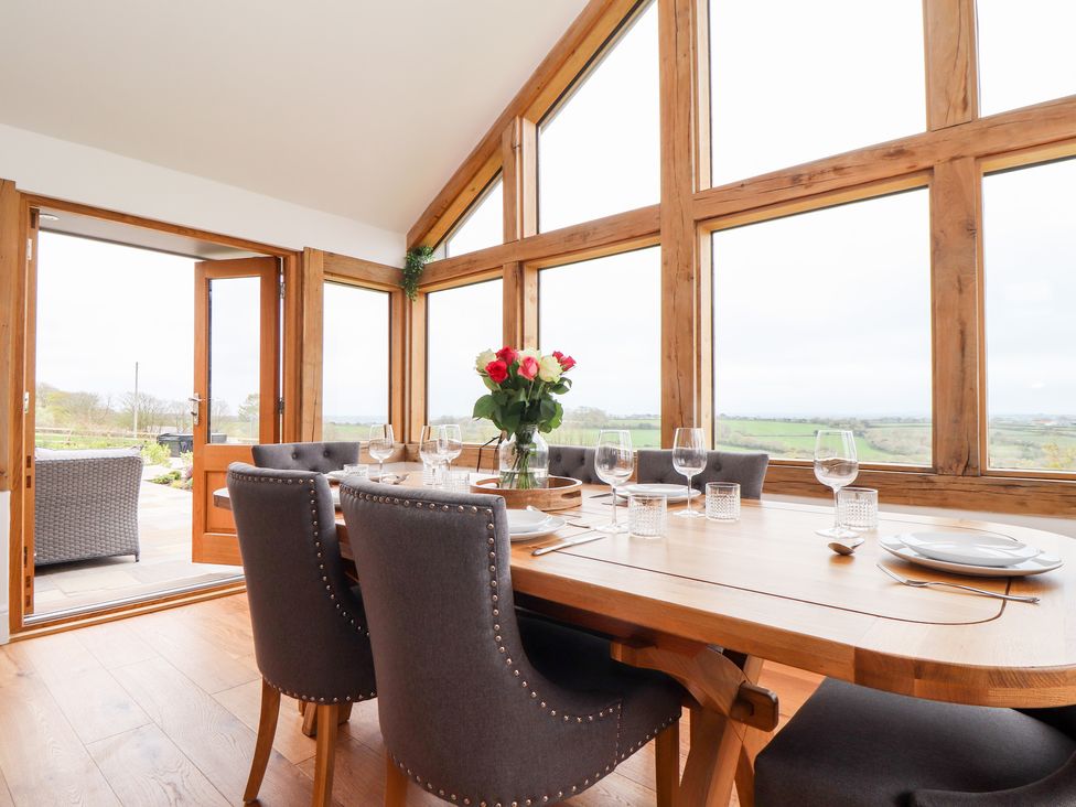 A dining room with a wooden table and grey chairs near large windows overlooking green fields at Trevilges Cottage in Trewennack near Helston