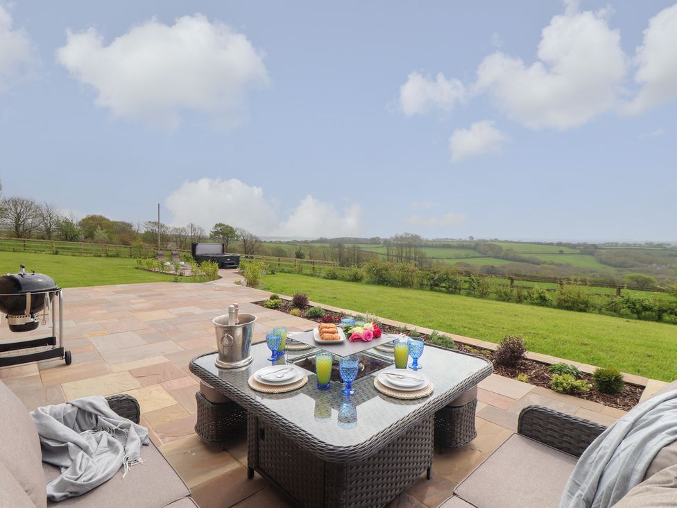 An outdoor patio area with a wicker dining table set with plates and glasses a grassy garden and countryside view at Trevilges Cottage in Trewennack near Helston