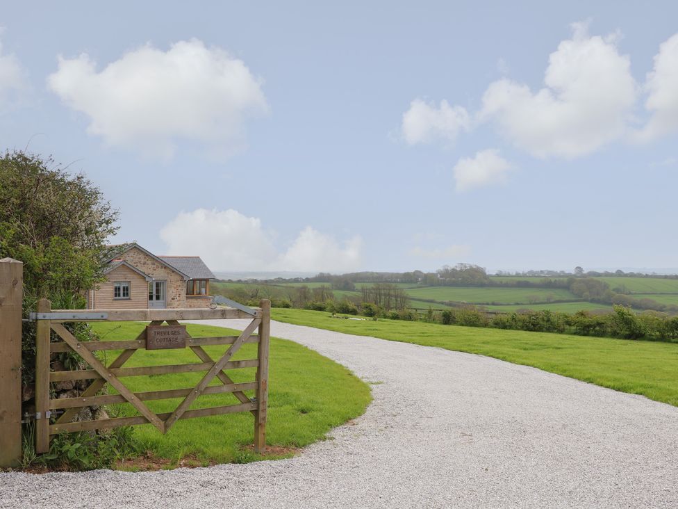 A gravel driveway with a wooden gate and a stone cottage surrounded by green fields at Trevilges Cottage in Trewennack near Helston