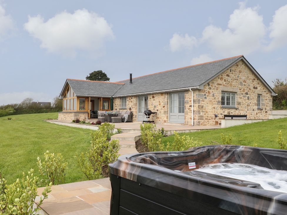 A stone house with a tiled roof and outdoor seating area with a hot tub and green lawn at Trevilges Cottage in Trewennack near Helston