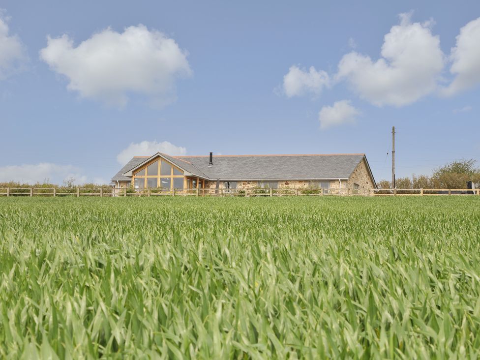 A stone house with large windows behind a fenced green field under a blue sky at Trevilges Cottage in Trewennack near Helston