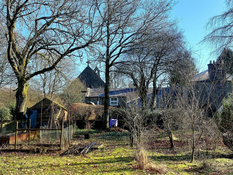 A garden with bare trees a wooden shed and stone buildings in the background at The Coach House Canonbie near Langholm