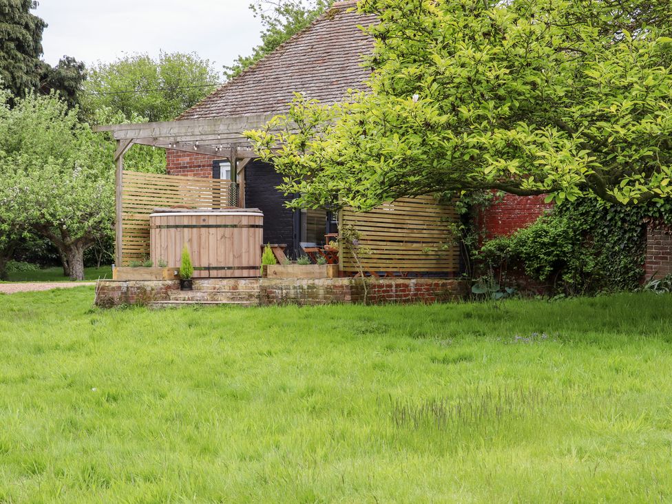 A grassy garden with trees and a wooden hot tub on a brick patio at The Snug at Pickelden Farmhouse in Mystole near Chilham