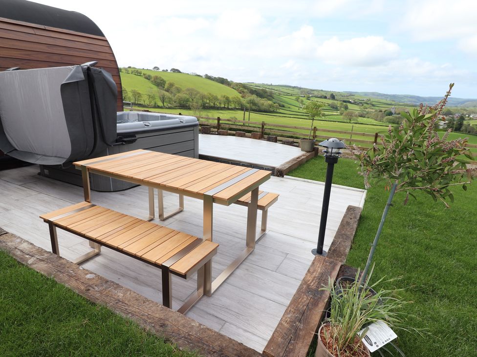 An outdoor patio with a wooden table and benches a hot tub a small tree and countryside views at Ceunant in Llanfihangel-y-Creuddyn near Aberystwyth