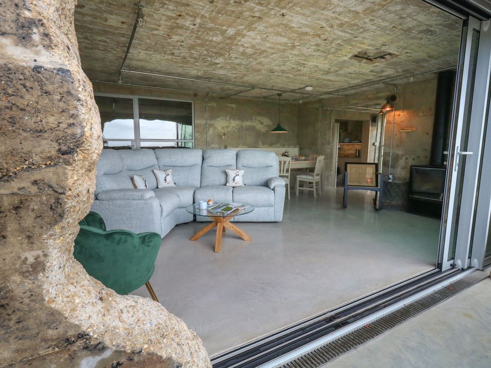 A living area with a gray sectional sofa and a glass coffee table next to a green chair by a large open sliding door at The Transmitter Bunker in Ringstead