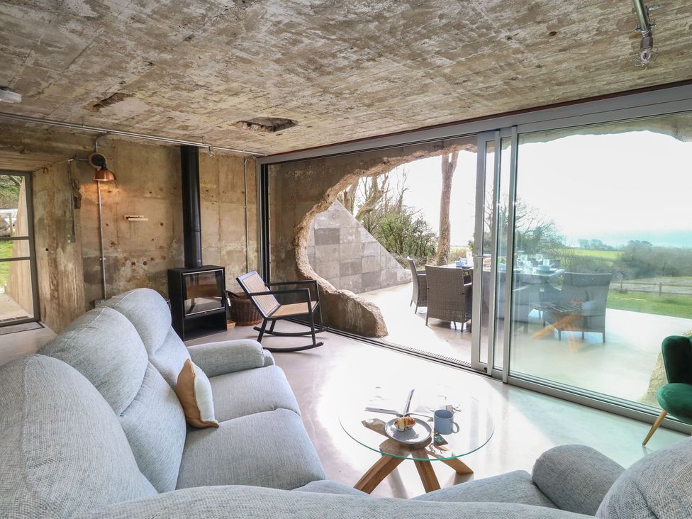 A living room with a gray sofa wooden rocking chair glass coffee table and sliding glass doors opening to an outdoor dining area at The Transmitter Bunker in Ringstead