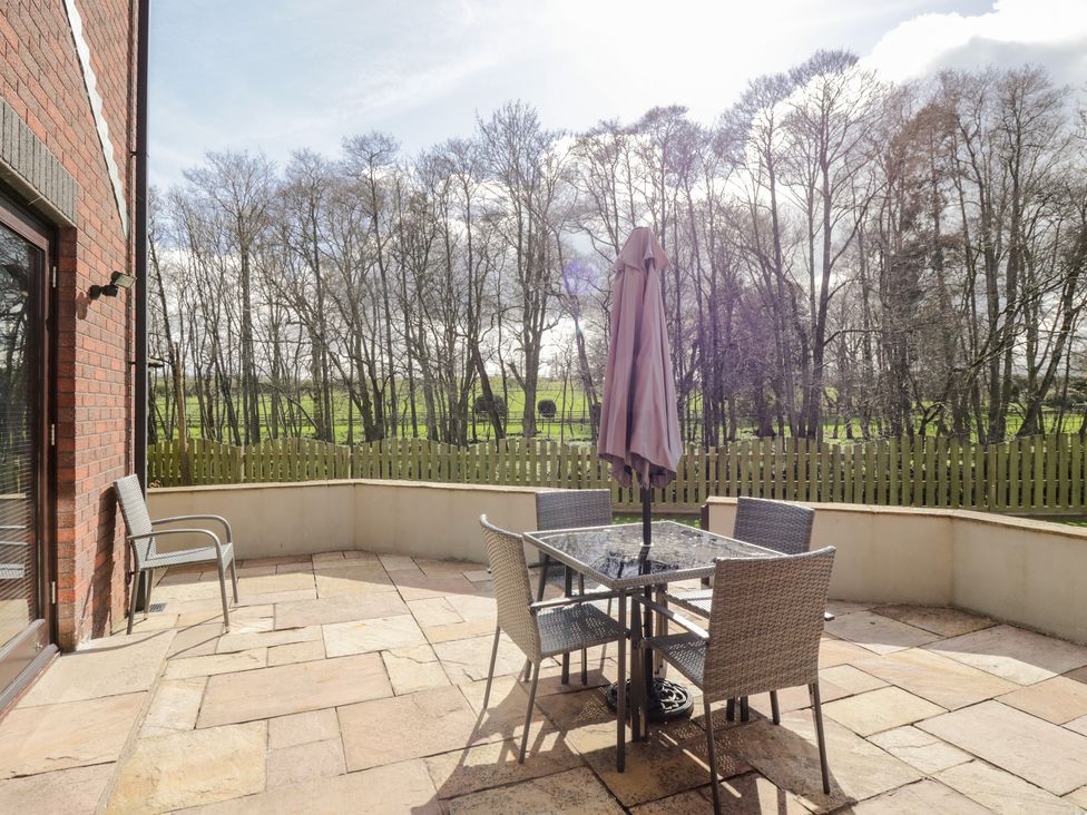 An outdoor patio with a table and four chairs and a closed umbrella surrounded by a low wall and a wooden fence with trees in the background at Mill End in Eamont Bridge near Penrith