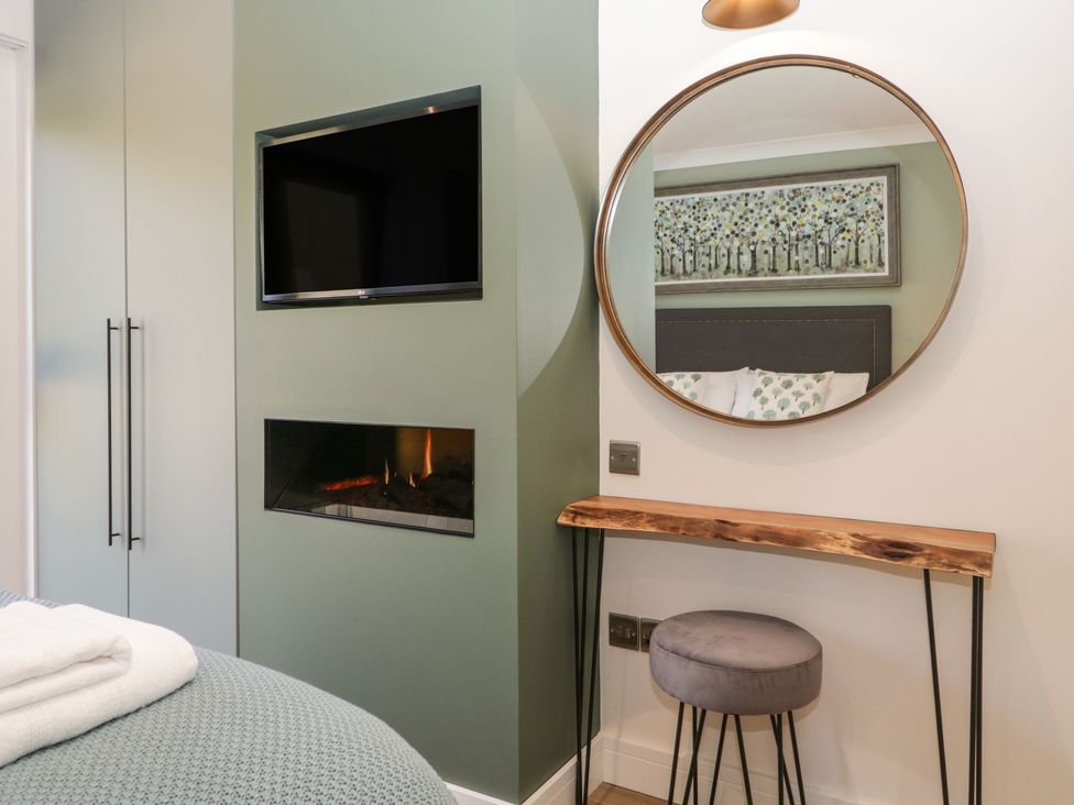 A bedroom with a wall-mounted television fireplace and a wooden console table with a round mirror and stool at Watch Tree Lodge in Allithwaite near Cartmel