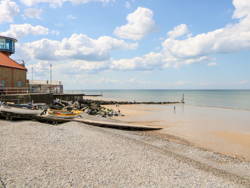 A beach with pebble shoreboats on a ramp and people on sand near a stone building at Beck Cottage in Sheringham