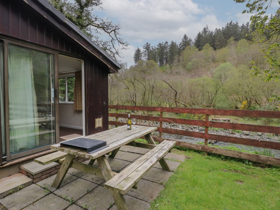 An outdoor wooden picnic table with a bottle and glasses on a patio next to a dark wooden cabin surrounded by trees and a river at Beech Timber Lodge Keswick