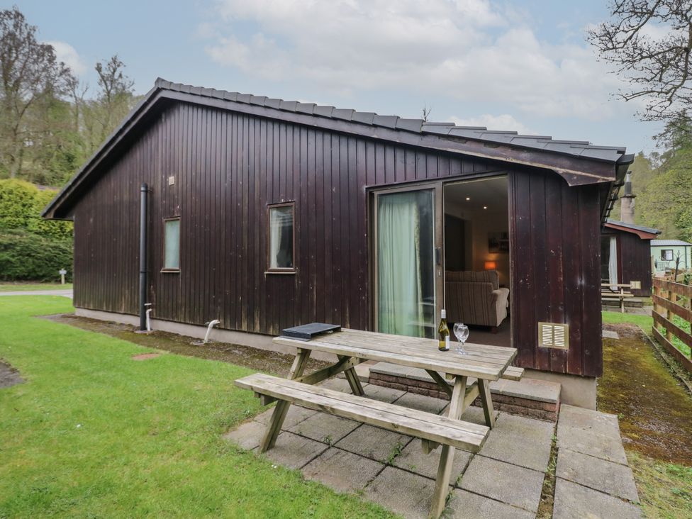 An outdoor wooden picnic table with a bottle and two glasses beside a dark wooden cabin with sliding glass doors at Beech Timber Lodge in Keswick