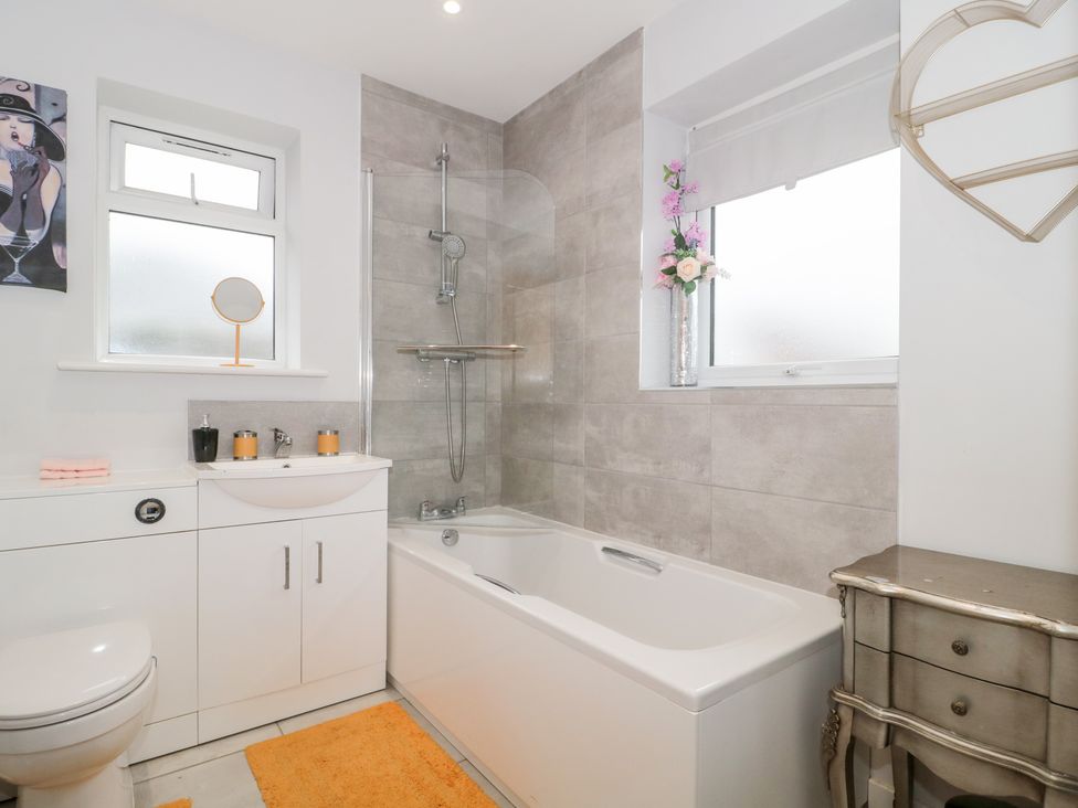A bathroom with a bathtub shower combination a toilet a white vanity unit with sink and a small cabinet with flowers at Paradise Villa in Highcliffe