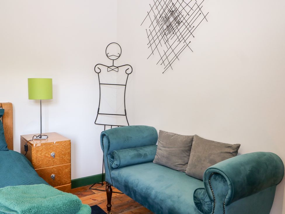 A bedroom corner with a teal chaise lounge sofa two gray pillows a green table lamp on a wooden nightstand and metal wall art at The Masters House Albourne near Hurstpierpoint