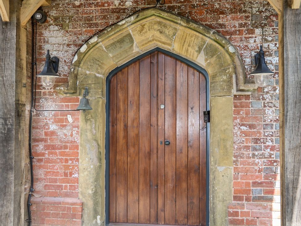 A wooden arched door set in a brick wall with stone trim and two wall-mounted lamps at The Masters House in Albourne near Hurstpierpoint