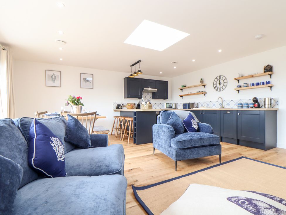 A living room and kitchen area with blue sofas a dining table with flowers and a kitchen with dark cabinets at Chi an Mor Lower Kenneggy near Praa Sands
