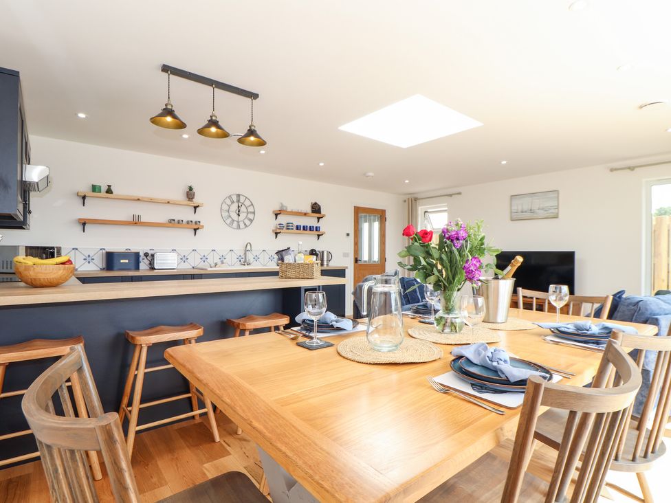 A dining area with a wooden table set with plates and glasses next to kitchen with bar stools and shelves at Chi an Mor in Lower Kenneggy near Praa Sands