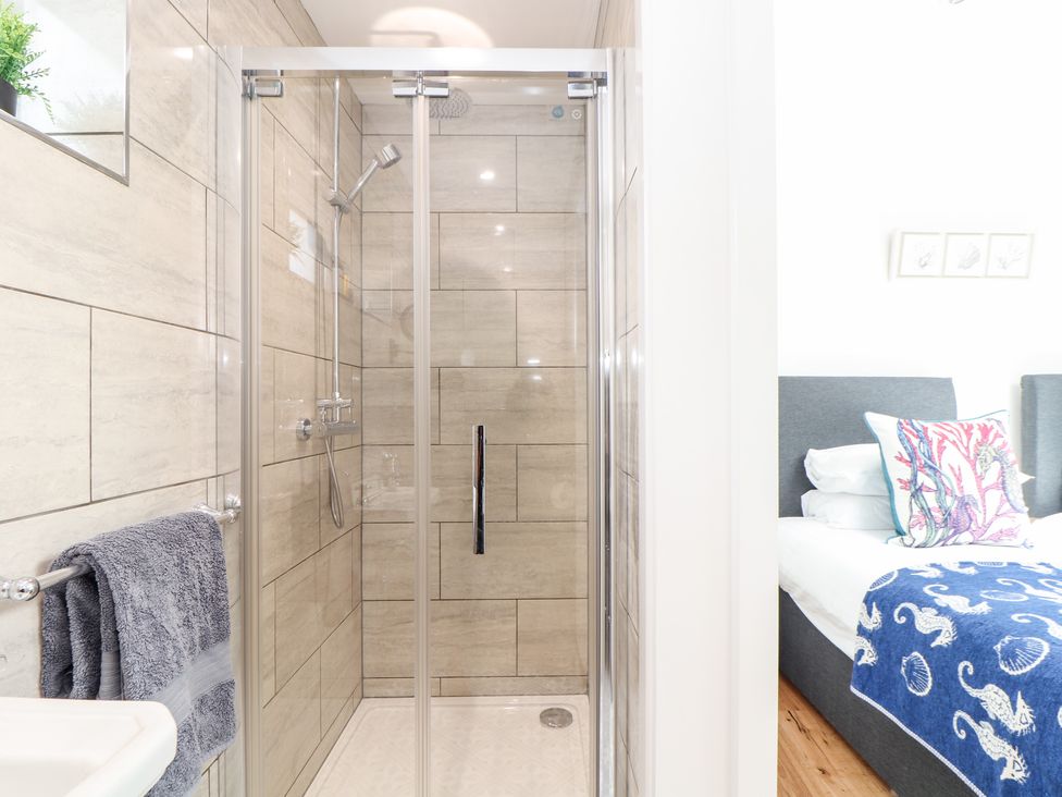 A bathroom with a glass shower enclosure tiled walls and a towel rack next to a bedroom with a bed and patterned cushions at Chi an Mor Lower Kenneggy near Praa Sands