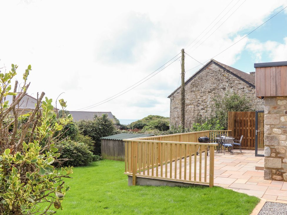 A garden with a grassy lawn and a wooden fenced patio with outdoor furniture and a stone building at Chi an Mor Lower Kenneggy near Praa Sands