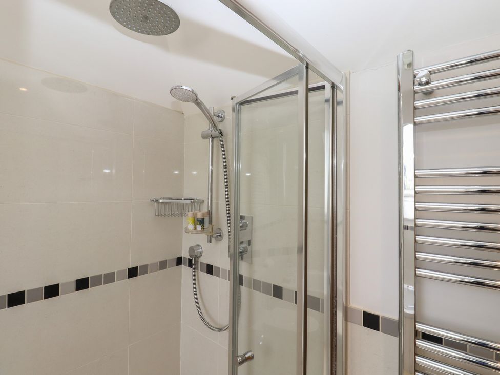 A bathroom shower area with a glass door a rainfall showerhead a handheld showerhead a wall-mounted shower caddy and a chrome towel radiator at Spindlewood Cottage in Hawkhurst