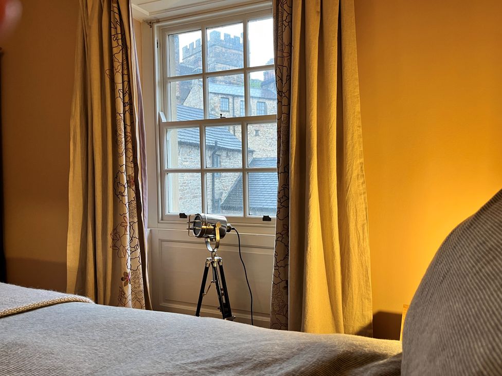 A bedroom window with patterned curtains and a spotlight lamp on a tripod with stone building visible outside at 5 Castle Hill in Lancaster