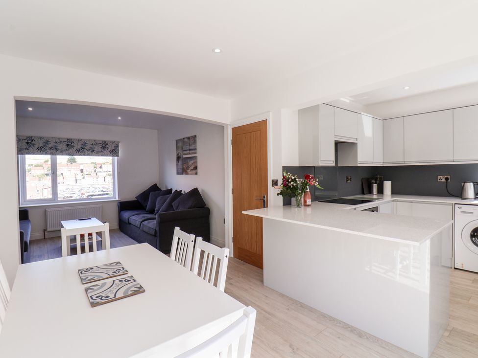 A kitchen with white cabinets and an island next to a dining area with a table and chairs and a living room with a sofa and a window at Whitby View in Whitby