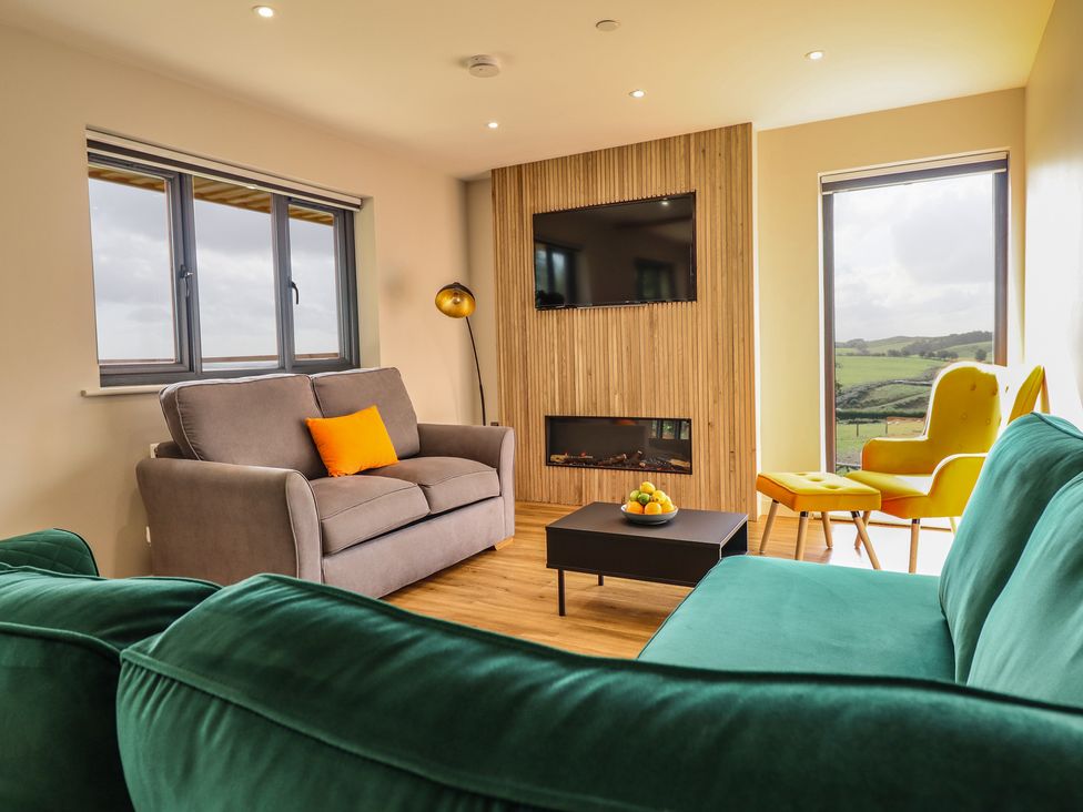 A living room with two sofas a yellow armchair and ottoman a TV mounted on wood paneling and a coffee table with fruit at Mynydd Y Glyn Lodge in Llanfyllin near Llanrhaeadr-Ym-Mochnant