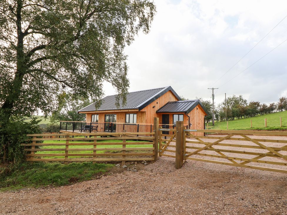 A wooden lodge with a metal roof behind a wooden gate with a gravel driveway and green fields at Mynydd Y Glyn Lodge Llanfyllin near Llanrhaeadr Ym Mochnant