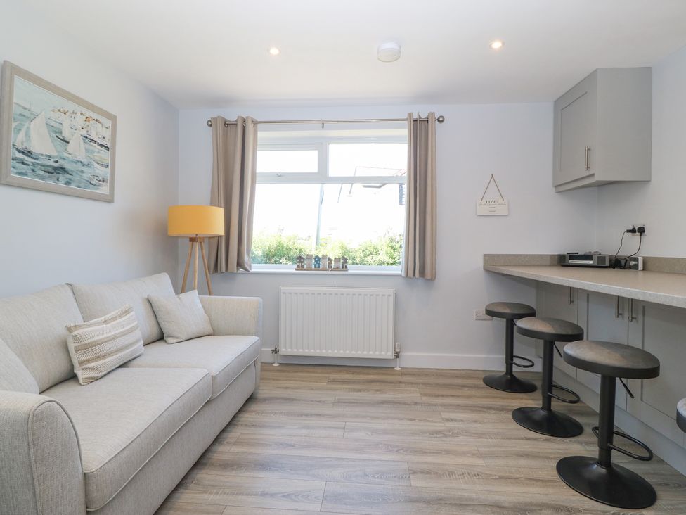 A room with a sofa pillows floor lamp window with curtains radiator and a kitchen counter with stools at Llwyn Eithin in Benllech