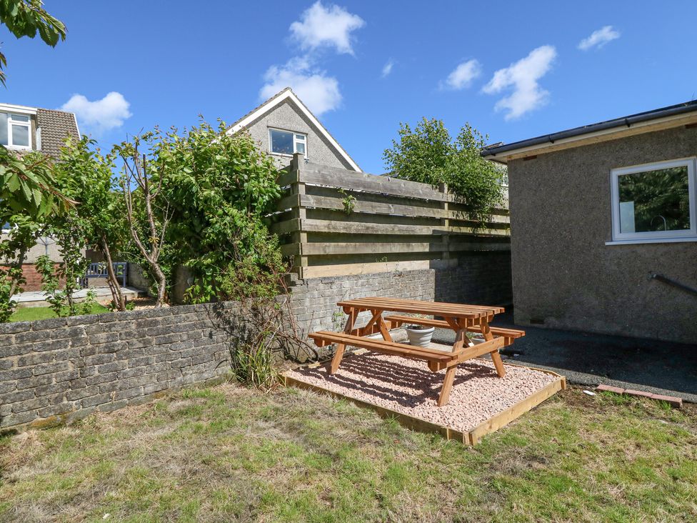 A wooden picnic table on gravel in a fenced backyard with grass and trees at Llwyn Eithin in Benllech