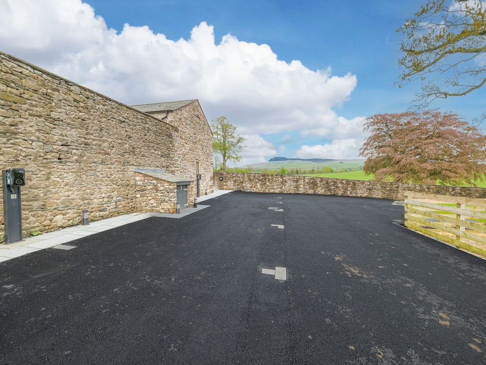 An empty paved parking area next to a stone building with a stone wall and distant hills at No 2 The Coach House in Thornton In Lonsdale