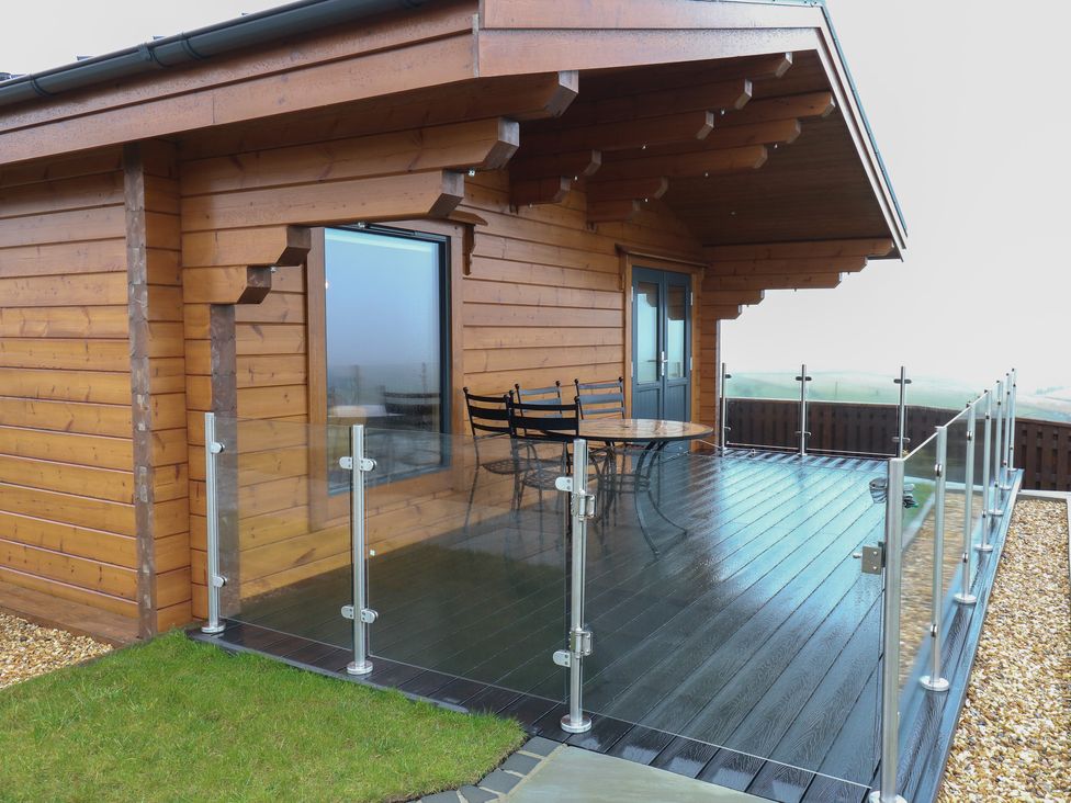 A wooden cabin with a glass railing and outdoor table with chairs on the deck at Height End Farm Log Cabin in Rossendale near Bacup