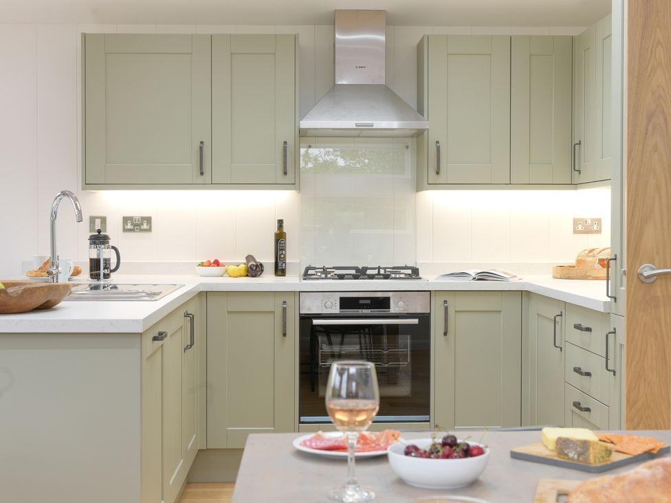 A kitchen with green cabinets white countertops a stove a sink a glass of wine and food on the counter at 4 Valley View in Lanreath