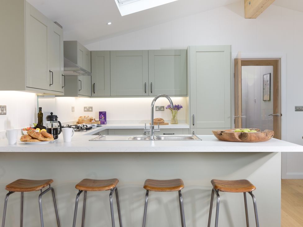 A kitchen with a countertop island with four stools a sink and faucet croissants and mugs on the counter and cabinets with under cabinet lighting at 4 Valley View in Lanreath