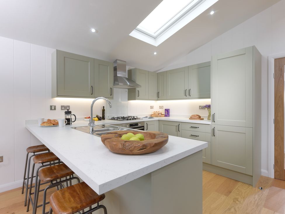 A kitchen with a white countertop island, wooden stools, green cabinets, a stainless steel sink, oven, gas stove, and a skylight window at 4 Valley View in Lanreath