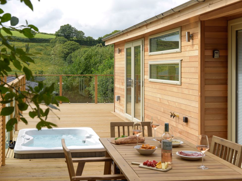 An outdoor wooden deck with a hot tub and a dining table set with food and drinks at 4 Valley View in Lanreath