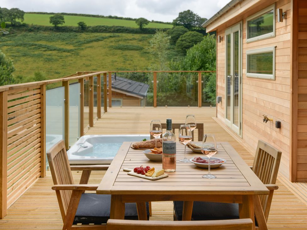 A wooden deck with a dining table set with wine glasses snacks and a bottle overlooking green hills at 4 Valley View in Lanreath