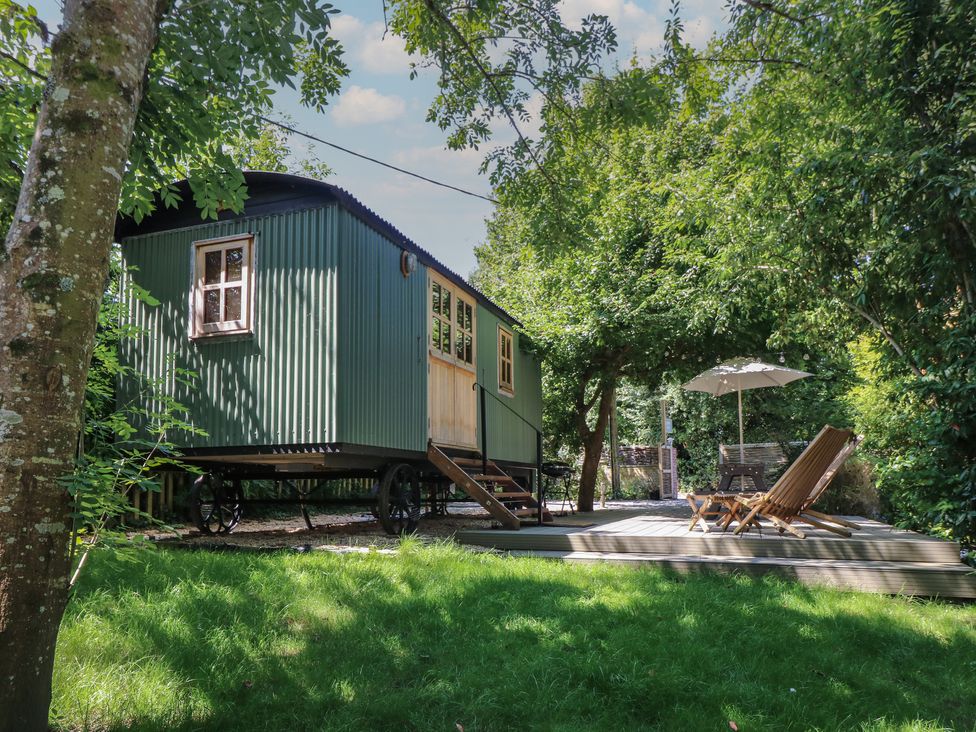 A green outdoor cabin on wheels with stairs leading to a wooden deck with chairs and an umbrella surrounded by trees at The Happy Hut in Sopley near Christchurch