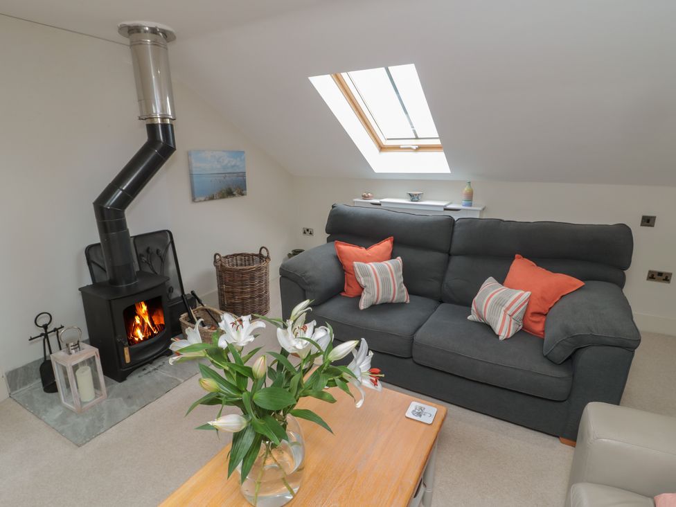 A living room with a dark grey sofa with orange and striped cushions a wooden coffee table with a vase of white flowers a wood burning stove and a skylight window at The Coach House in Bamburgh