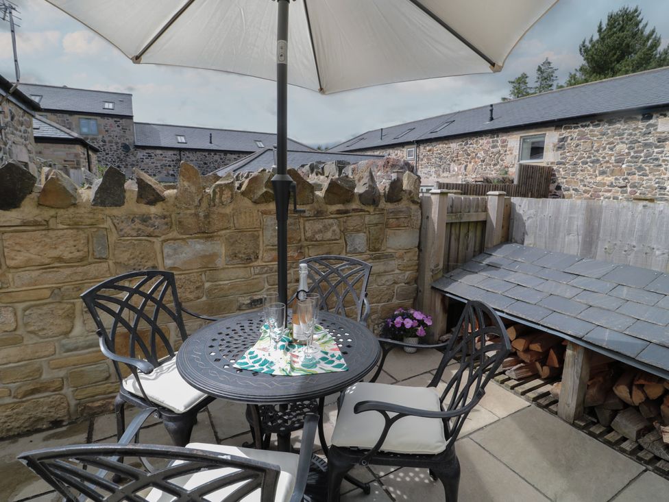 An outdoor patio with a round metal table and four chairs under a large umbrella near a stone wall and wood storage at The Coach House in Bamburgh